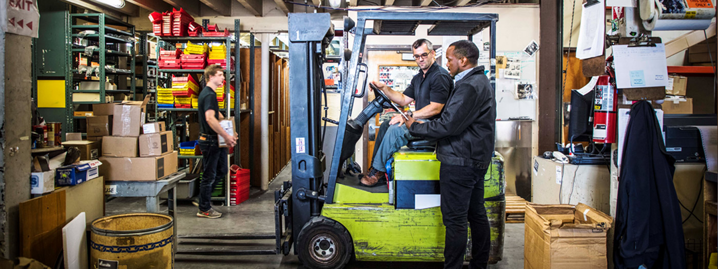 Trois employés travaillant dans l'entrepôt. Un homme est debout et utilise une tablette, parlant avec un autre employé assis dans un chariot élévateur. Un troisième homme est à l'arrière-plan portant une boîte en carton.