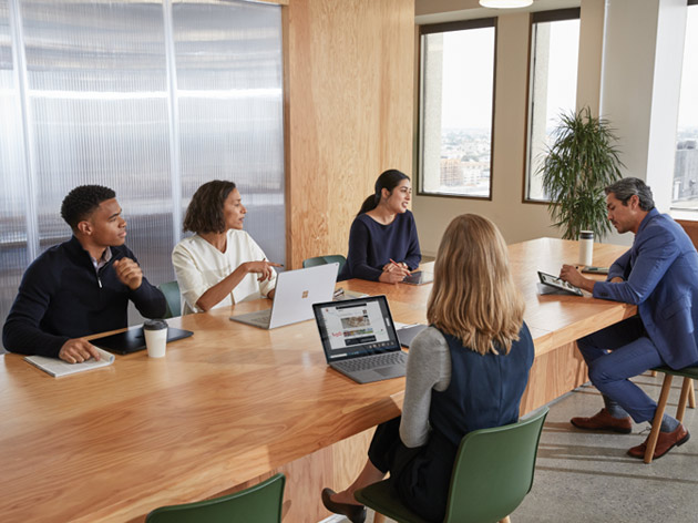 Cinq adultes collaborant dans une salle de conférence, tous assis à une table de conférence à l'aide d'appareils du portefeuille Microsoft Surface. Deux écrans sont visibles avec le tableau blanc Microsoft affiché.