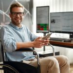 Successful trader. Portrait of young and cheerful male businessman in a wheelchair holding his smartphone and smiling while sitting at his workplace in modern office. Disabled people