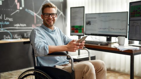 Successful trader. Portrait of young and cheerful male businessman in a wheelchair holding his smartphone and smiling while sitting at his workplace in modern office. Disabled people