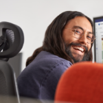 Man smiling while at work​ Keywords: Viva; Filip; hybrid work; work from home; remote; male; desktop monitor; UI screen; glasses; looking back over his shoulder; office; ; Insights; praise