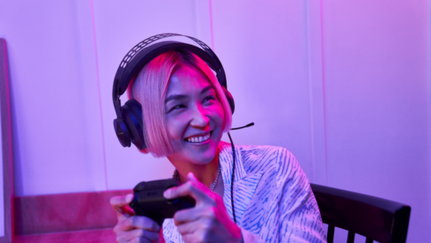 Contextual image of Gen Z woman inside home gaming on laptop with headset and controller