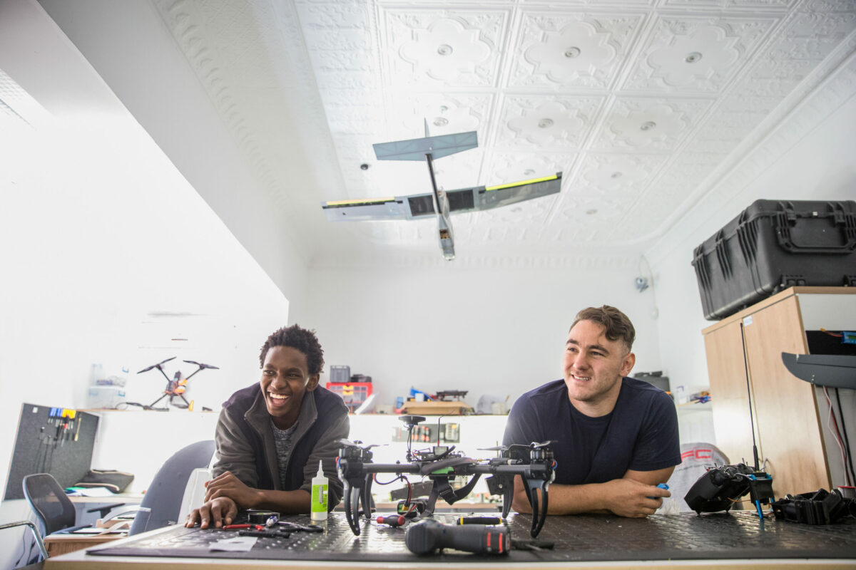Deux jeunes garçons souriants, assis dans un open space, entourés de drones et de matériel de fabrication de drones.