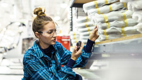 Travailleuse portant des lunettes de protection, agenouillée devant des étagères en métal dans un espace de stockage commercial de fabrication. Elle tient un appareil IIoT Askey.