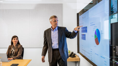 Homme d'affaires en costume faisant une présentation dans une salle de conférence de bureau. Il pointe un grand écran de moniteur affichant un graphique circulaire et plusieurs statistiques/valeurs monétaires.